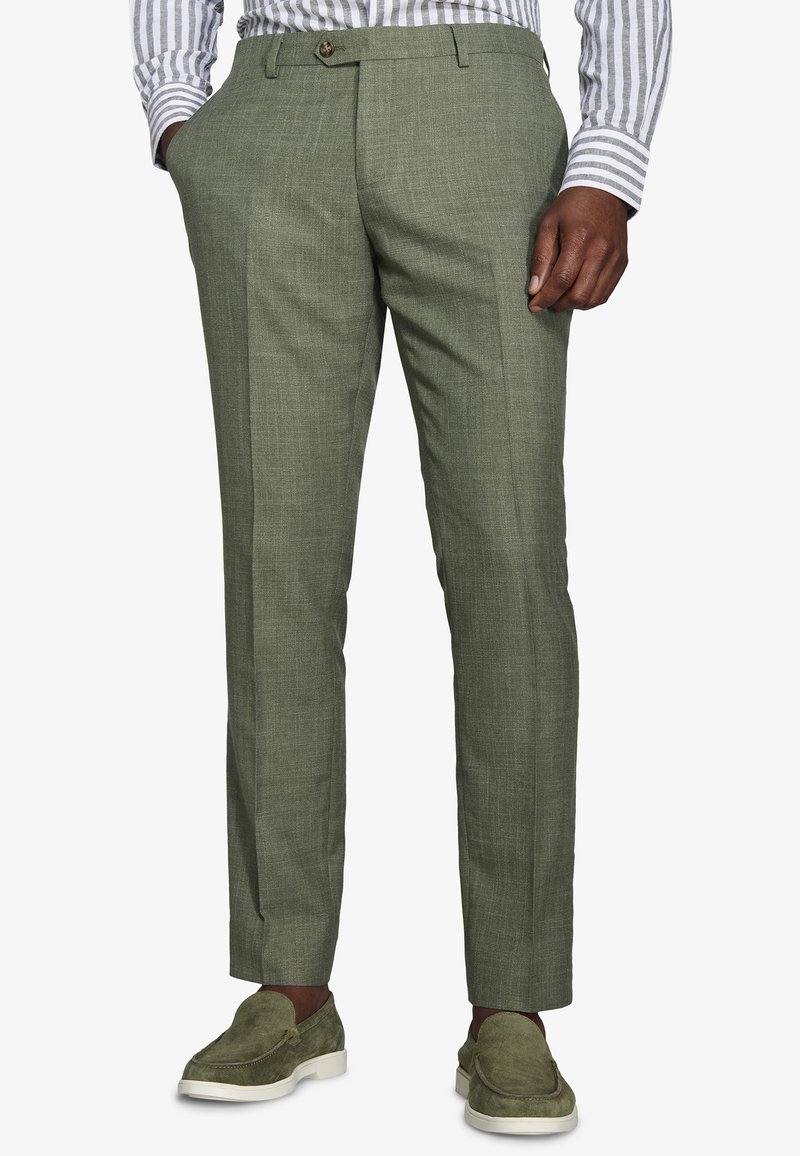 Hombre con pantalones a medida de color verde oliva, camisa a rayas y mocasines de ante verde oliva a juego con suelas blancas, de pie con una mano en el bolsillo.