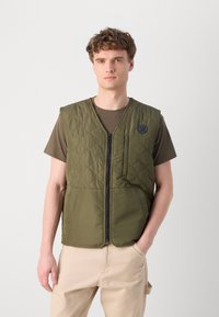 Olive green quilted vest with a front zipper, two pockets, and a small logo patch on the left chest; worn over a brown t-shirt.