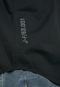 Černé tričko s kulatým lemem, s šedým potištěným textem "J-F/63.001" na boku, vyrobené z hladké tkaniny.