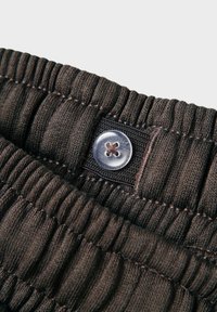 Elastīga josta tumši brūnā audumā ar horizontālu rievotu tekstūru. Iespējams, caurspīdīgs pogas ar dekoratīvu zvaigznes formu.