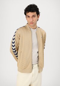 Beige bomber jacket with tonal floral pattern, navy striped sleeves, ribbed cuffs, and front zipper. Paired with a light gray shirt.