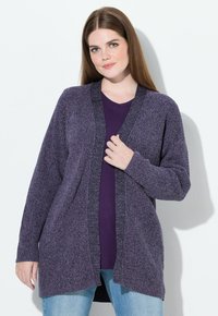 Kobieta z długimi brązowymi włosami, ubrana w fioletowy, dziergany kardigan na ciemnofioletowej koszuli i jasnoniebieskich dżinsach, stojąca na białym tle.