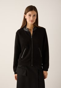 Schwarze Bomberjacke mit Reißverschluss, strukturiertem Stoff, Lederakzenten am Kragen und Seitentaschen. Wird über einem schwarzen Rock getragen.