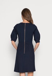 Mörkblå klänning med puffärmar, figurnära överdel och A-linjeformad kjol. Har en guldfärgad dragkedja på baksidan och en mjuk tygkvalitet.