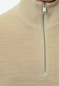 Pull en tricot côtelé beige avec une demi-fermeture éclair argentée et un col montant, montrant la partie supérieure de la poitrine et du cou.