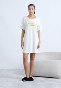 Λευκό νυχτικό με κοντά μανίκια, με το γράφημα "Φτιάξε λεμονάδα!" σε κίτρινα και μαύρα γράμματα. Φορεμένο με μαύρες χνουδωτές παντόφλες.