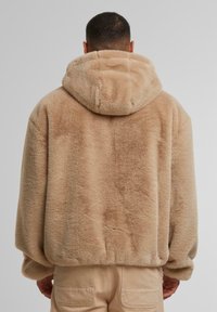Hellbraune Kunstfell-Hoodie mit weicher Textur, großen Kapuze und lockerer Passform. Von hinten betrachtet hat es ein sauberes, minimalistisches Design.