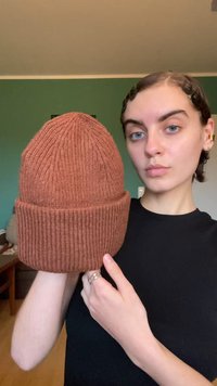 Brown knitted beanie with vertical ribbed texture, featuring a folded cuff. Presented by a person against a green wall background.