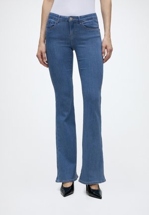 Vrouw draagt mid-rise blauwe bootcut jeans en zwarte puntige hakken, staand tegen een effen witte achtergrond.