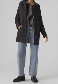 Abrigo negro resistente al clima con capucha, combinado con una camisa a rayas y jeans de pierna ancha en azul claro. El conjunto se completa con zapatos de plataforma negros.