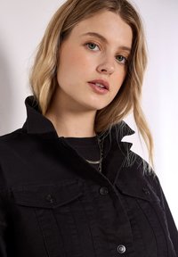 Czarna kurtka dżinsowa z kołnierzem, dwoma kieszeniami na piersi zapinanymi na guziki, pionowymi przeszyciami oraz luźnym fasonem. Nadaje się do noszenia na co dzień.