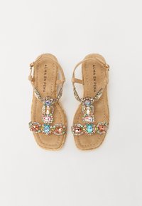 Sandali in suede beige con gemme decorative multicolori, dotati di un design a T e una suola piatta con dettagli intrecciati.