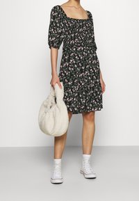 Schwarzes Blumen Kleid mit kurzen Puffärmeln, gerafftem Oberteil und gerafftem Saum, kombiniert mit einer cremigen plüschigen Tragetasche und weißen High-Top-Sneakern.