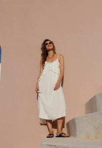 Robe blanche sans manches avec des accents volants, longueur mi-mollet. Le modèle se tient sur des marches en portant des sandales noires, devant un mur rose.