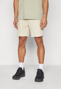 Beige shorts med et struktureret stof, lidt over knæene. Matchet med sorte træningssko og hvide ankelsokker. Casual pullover-top.
