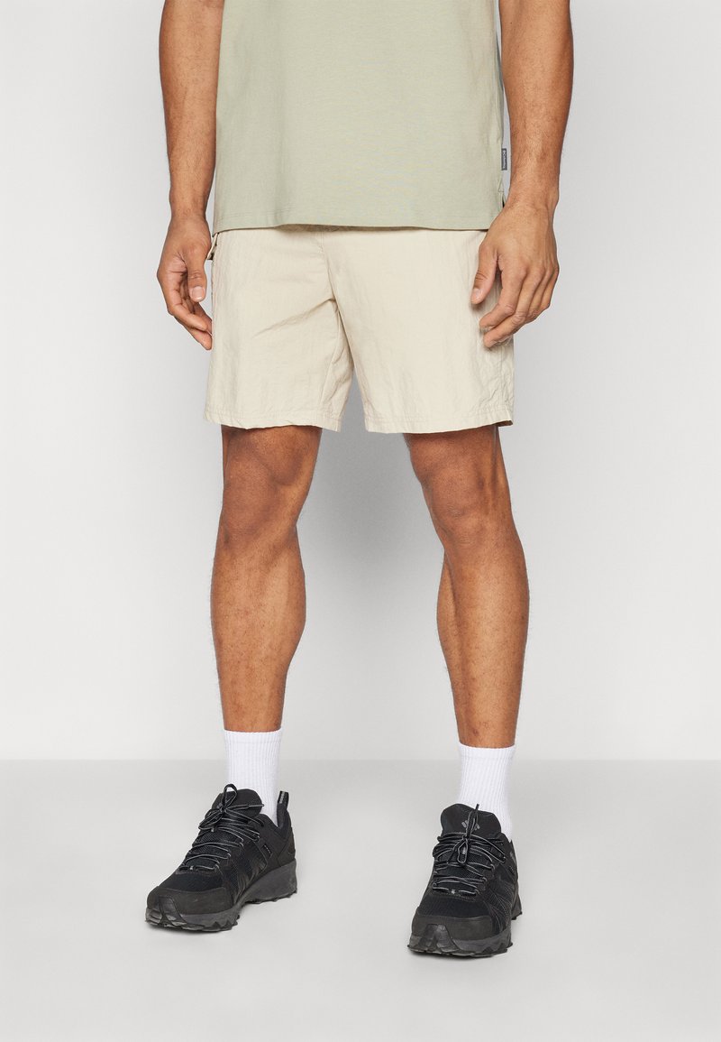 Beige shorts med et struktureret stof, lidt over knæene. Matchet med sorte træningssko og hvide ankelsokker. Casual pullover-top.