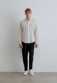 Baltas dryžuotas marškinėlis ilgomis rankovėmis su atsegamu apykakliu, derinamas su juodomis kelnėmis ir baltais sportbačiais. Paprastas dizainas, minimalūs akcentai.