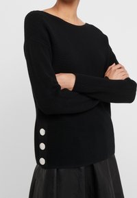 Un pull noir en tricot avec une texture côtelée, un large col, et trois boutons décoratifs en argent le long du côté gauche.