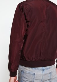 Burgunderfarbene Bomberjacke mit glatter Textur und gerippten Bündchen, ausgestattet mit einem taillierten Saum und schlichtem Design. Keine sichtbaren Beschläge oder Akzente.