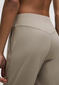 Nahaufnahme einer Person, die hochgeschnittene beigen Leggings trägt, die sichtbare Seitennähte und ein kleines Logo am Bund aufweisen.