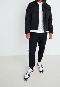 Svart vadderad jacka med dragkedja fram, vit t-shirt, svarta slim-fit byxor, och svarta/röda sneakers med vit sula. Avslappnad outfit.