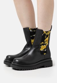 Botas de tobillo de cuero negro con suelas gruesas, paneles laterales elásticos que presentan un patrón ornamental en amarillo y dorado, llevadas sobre piernas desnudas.
