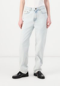 Jeans de denim azul claro con corte recto, cintura alta, costuras visibles, dos bolsillos frontales y cierre de botón. Combinados con mocasines negros.
