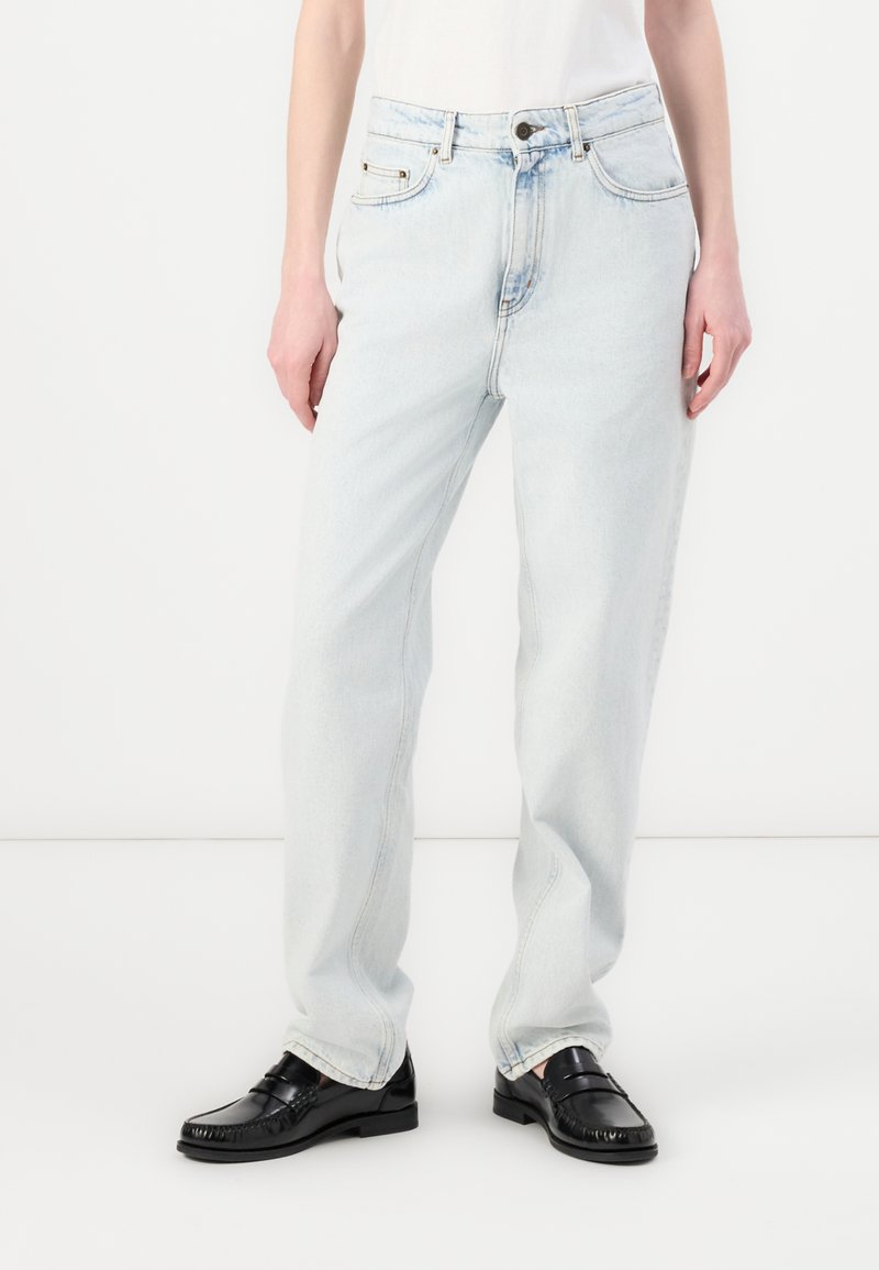 Jeans de denim azul claro con corte recto, cintura alta, costuras visibles, dos bolsillos frontales y cierre de botón. Combinados con mocasines negros.