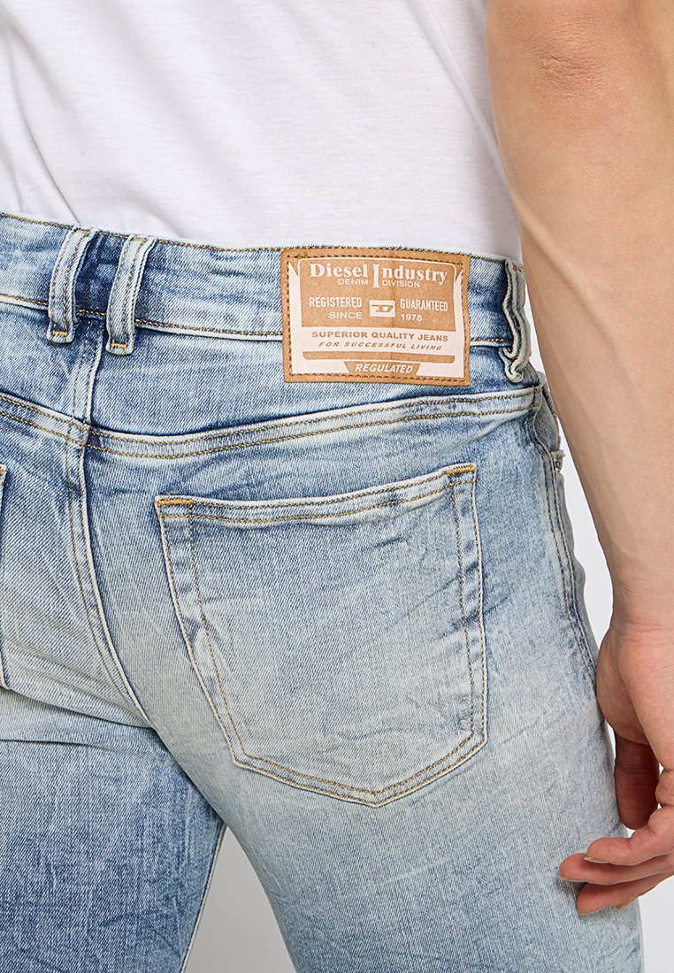 Jeans in denim blu sbiadito con un'etichetta in pelle color carne che presenta il marchio "Diesel Industry". Design a cinque tasche con dettagli di cucitura visibili.