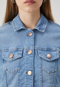 Chaqueta de mezclilla en azul claro, con cuello extendido y cuatro bolsillos frontales. Los botones de tono cobre con detalles grabados complementan el diseño.
