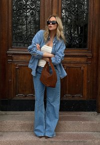 Džinsu krekls, balta caurspīdīga blūze, augstās jostasvietas zilie džinsi, brūna zamša rokassoma, melni saulesbrilles; stāv uz akmens kāpnēm pretī koka durvīm.