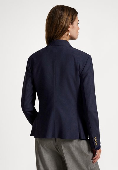 Kobieta widziana od tyłu, ubrana w granatową marynarkę z złotymi guzikami na rękawie oraz szare spodnie, na jednolitym tle.