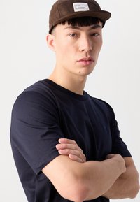 Tricou din bumbac bleu marin, cu guler rotund, asortat cu o pălărie din suede maro, având un mic etichetă rectangulară. Croiala este casual.