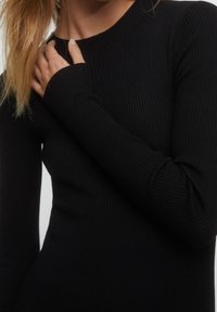 Haut noir à manches longues côtelé avec un col ras du cou. Le tissu a une finition lisse et texturée, avec des manches ajustées qui se rétrécissent aux poignets.