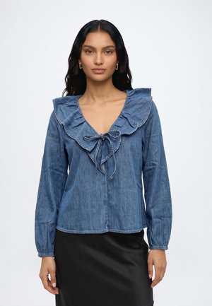 Femme aux longs cheveux foncés portant un chemisier en denim bleu avec un col à volants et un nœud, associé à une jupe noire, debout devant un fond uni.