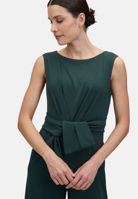 Zöld, ujjatlan ruha lekerekített nyakkivágással, csavart anyagrésszel a deréknál, és sima textúrával. Nincsenek látható minták vagy kiegészítők.