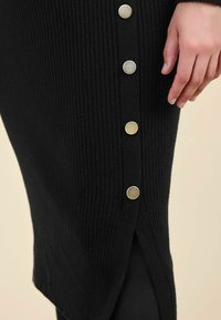 Abito nero a maglia con texture a righe verticali, spacco laterale e cinque bottoni metallici lungo la cucitura sinistra per un tocco di stile.
