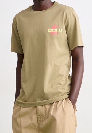 Camiseta de algodón verde oliva con un logo y gráfico en rojo y amarillo de "CONVERSE". Diseño de manga corta y cuello redondo. Textura suave.