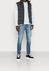 Gilet imbottito nero sopra una felpa azzurro chiaro, abbinato a jeans azzurri e sneakers nere. I dettagli includono una chiusura con zip e un design casual.