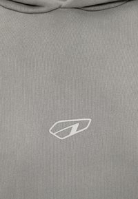 Γκρίζο υφασμάτινο ύφασμα με μικρό, λευκό αφηρημένο λογότυπο κεντραρισμένο κοντά στην κορυφή κάτω από μια μαζεμένη ραφή.