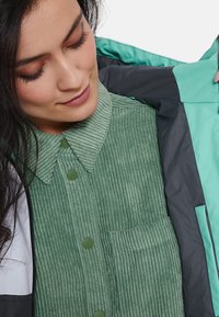 Žalias kordūroviškas marškinėliai su apykakle, turintys žalius mygtukus ir kišenę, dėvimi po teal ir pilkos spalvos švarku.