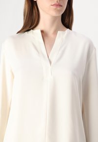 Blusa bianca sporca con scollo a V e maniche lunghe. Tessuto liscio, design semplice, con una piega centrale per dettagli.