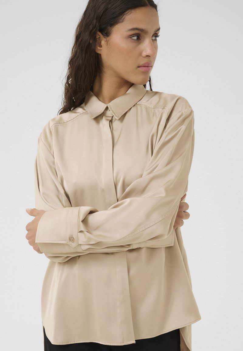 Beige satin skjorte med klassisk krave, knaplukning og lange ærmer. Har en løs pasform og rundet kant, som fremhæver den glatte tekstur.