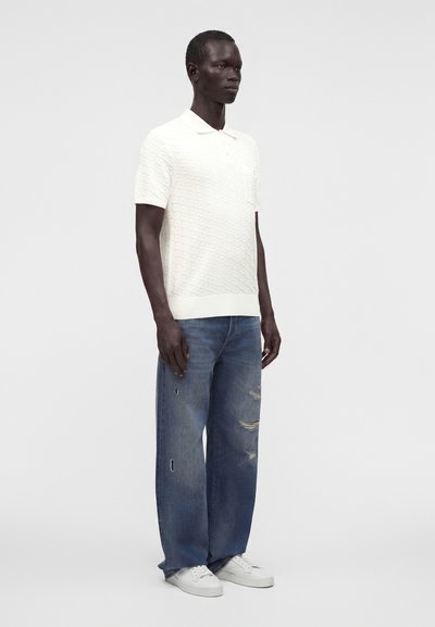 Homme portant un polo en maille blanc à manches courtes, un jean bleu délavé ample et des baskets blanches, debout devant un fond gris uni.
