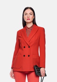 Roter, zweireihiger Blazer aus strukturiertem Stoff, mit zwei Fronttaschen und dekorativen schwarzen Knöpfen. Kombiniert mit einem gemusterten Hemd und einer schwarzen Handtasche.