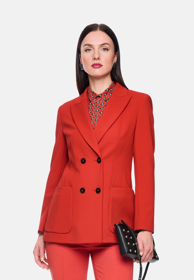 Roter, zweireihiger Blazer aus strukturiertem Stoff, mit zwei Fronttaschen und dekorativen schwarzen Knöpfen. Kombiniert mit einem gemusterten Hemd und einer schwarzen Handtasche.