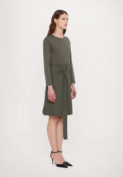 Abito in maglina verde oliva con maniche lunghe, vita con cintura dotata di un anello argentato e due tasche laterali; indossato con tacchi neri a punta.