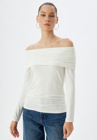 Off-the-shoulder witte top met gerimpelde stofdetails en lange mouwen, gecombineerd met lichtblauwe jeans. Soepele textuur, lichtgewicht materiaal.