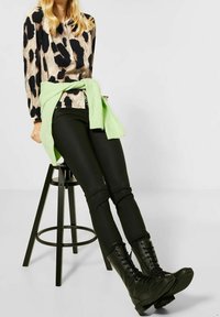 Stivali combat neri in pelle con tacco alto, pantaloni scuri aderenti e una maglietta a maniche lunghe con una stampa animalier beige e nera, con un maglione verde legato in vita.