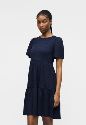 Femme avec des cheveux tressés portant une robe bleu marine longueur genoux avec des manches courtes bouffantes et un motif ajouré en zigzag.